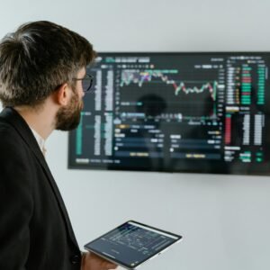 A businessman examines stock market data displayed on a monitor, holding a tablet.